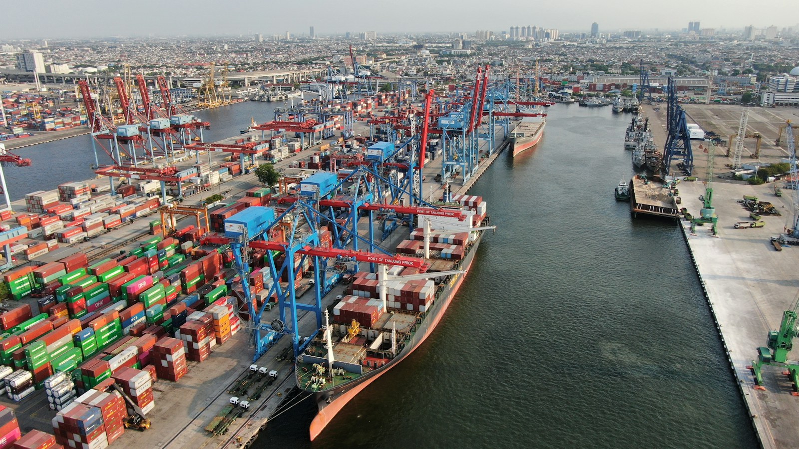 Aerial view of container ships, stacked cargo containers and cranes along a busy port channel.